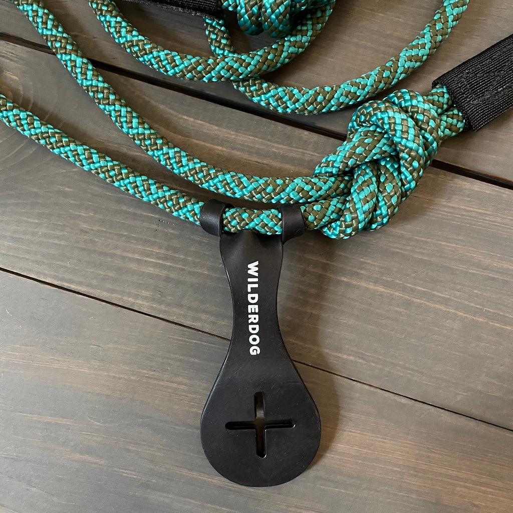 Image shows a teal and black climbing rope-style dog leash from Wilderdog, coiled with a plastic loop holder attached. The leash, laid out on a wooden surface, showcases its durable weave and the brand name prominently displayed on the holder. A hands-free Crap Carrier attachment ensures convenience during walks.