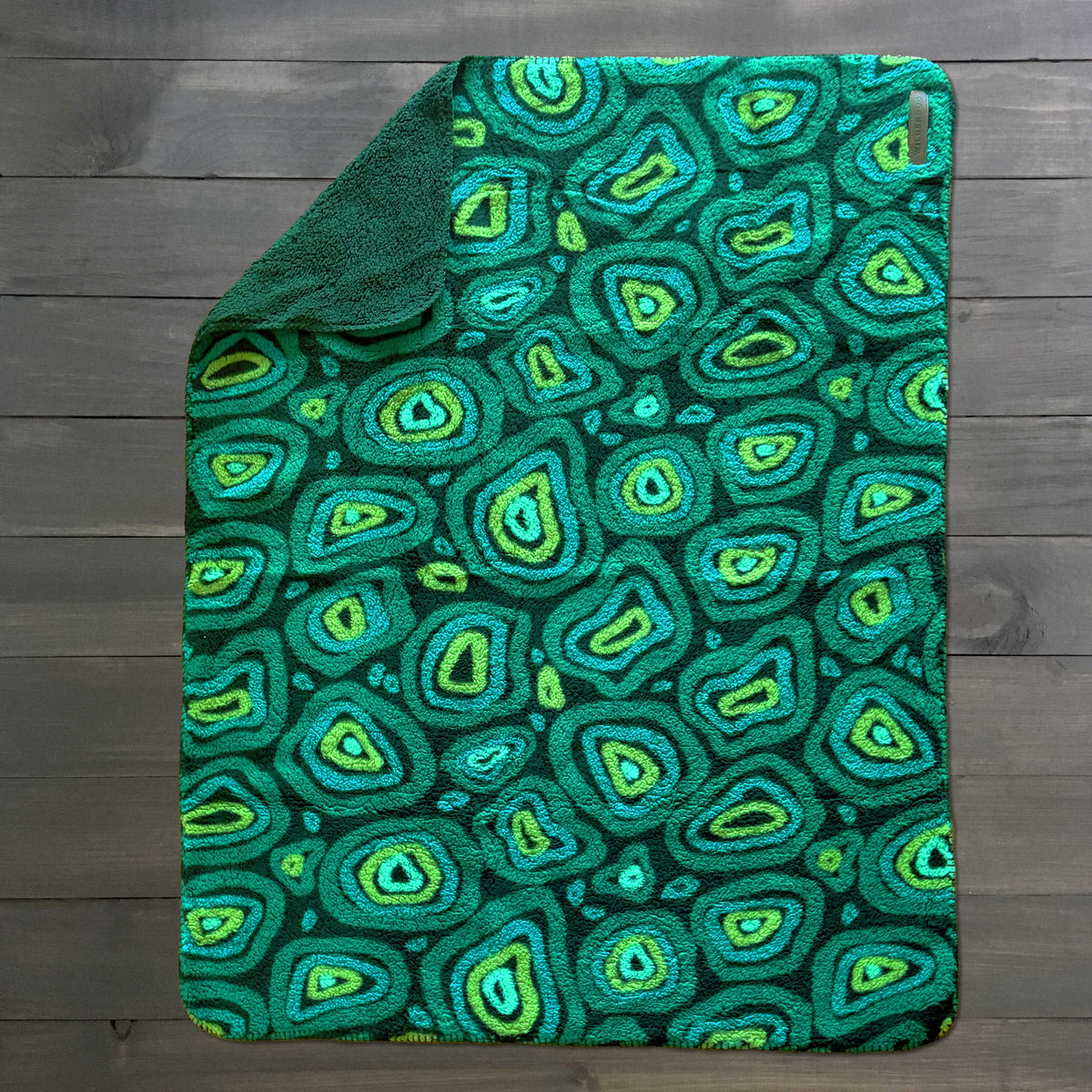 Sherpa Fleece Waterproof Blanket in green and blue, with a folded corner, placed on a dark wooden surface.