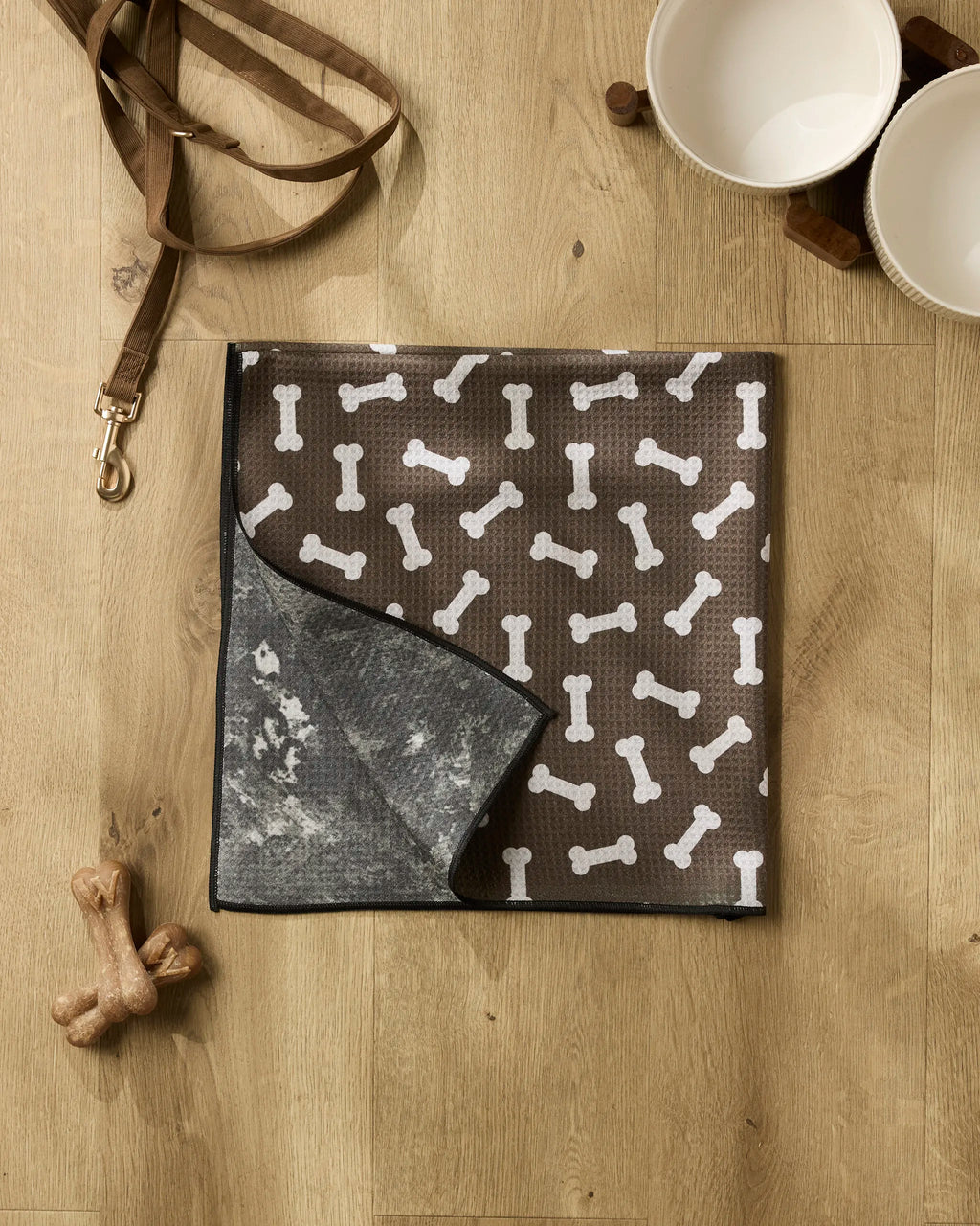 A brown dog mat with white bone patterns from Geometry's Sweet Treats collection lies on a wooden floor, joined by a recycled dog towel, leash, two ceramic bowls on a wooden stand, and two dog bone toys for a cozy corner.