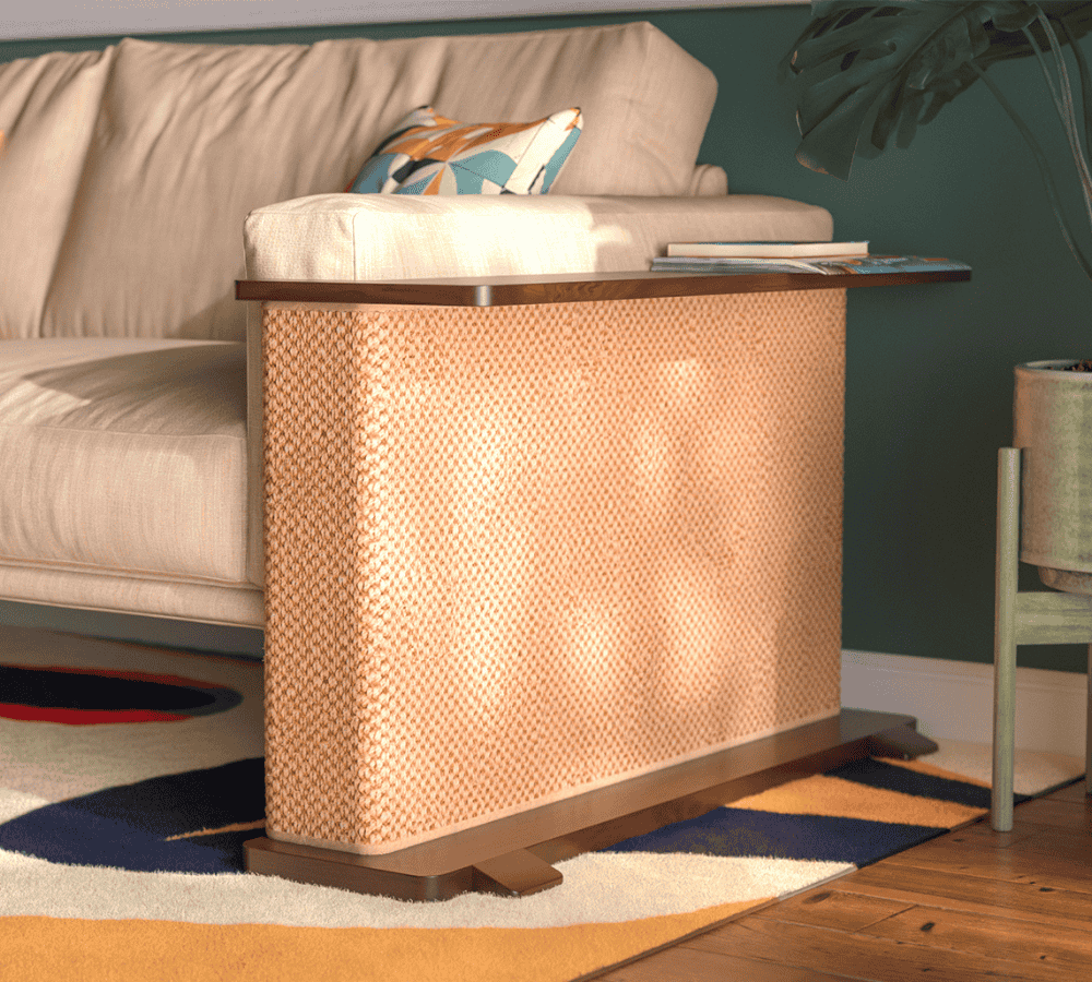 A Cat-e-Corner table placed next to a sofa, integrating a cat's scratching post with a small table, in a room with a plant and a colorful cushion.