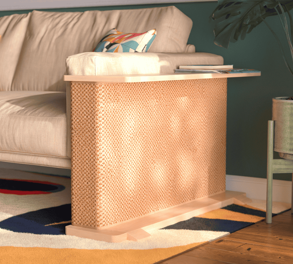 A Cat-e-Corner table placed next to a sofa, integrating a cat's scratching post with a small table, in a room with a plant and a colorful cushion.