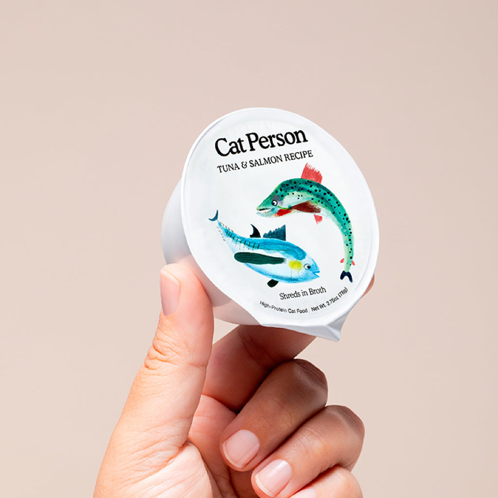 A hand holds a container of Cat Person Tuna & Salmon Shreds in Broth, featuring tuna and salmon illustrations on the lid. This protein-rich, balanced cat food is shown against a neutral background.